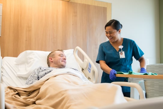 A smiling EVS team member in scrubs wipes down a surface while interacting with a patient in a hospital bed, creating a positive and clean patient environment.